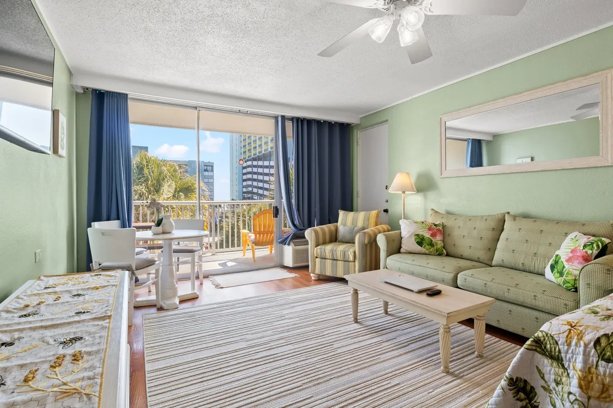 The image showcases a welcoming condo interior featuring a cheerful sofa with floral cushions, a sturdy wooden coffee table, and a private balcony offering a glimpse of the outdoors. Sunlight fills the space, highlighting a striped area rug and inviting guests to imagine relaxing mornings or cozy evenings with friends.