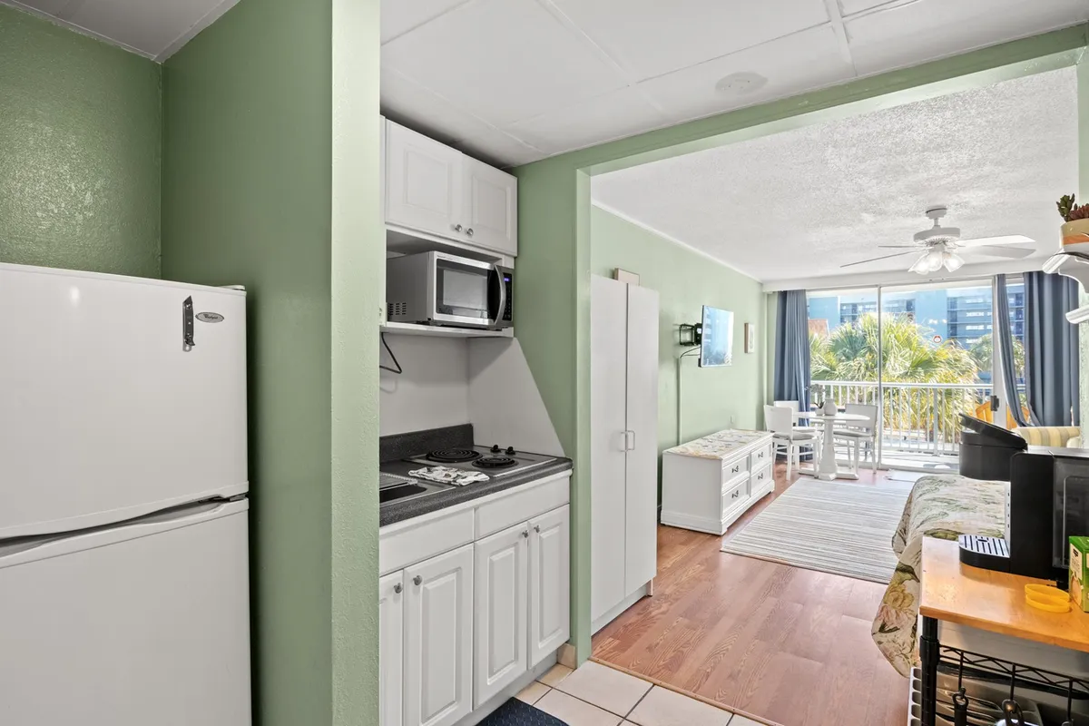 A bright interior scene featuring the kitchen and dining area with large windows, letting in natural light and offering glimpses outside. The space is set up for shared meals, morning coffee, and home-cooked comfort.