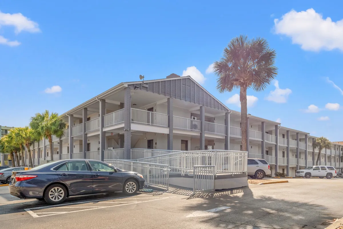 The exterior of a condo building framed by palm trees, visible balconies, and an inviting entrance ramp. Parked cars suggest convenient access. The scene hints at moments spent relaxing on balconies after a day at the beach.