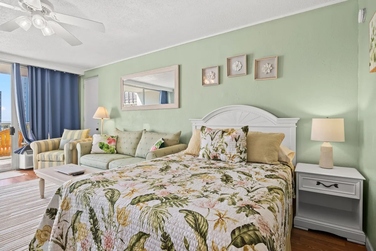 This image showcases the main sleeping area, featuring a bed adorned with a cheerful floral quilt and a sofa with decorative cushions. A large window lets plentiful natural light stream in, while gentle décor elements like wall art with shells and a decorative mirror add to the tranquil and inviting vibe. The arrangement highlights both the queen bed and the sleeper sofa, spotlighting the flexibility and comfort of the space.