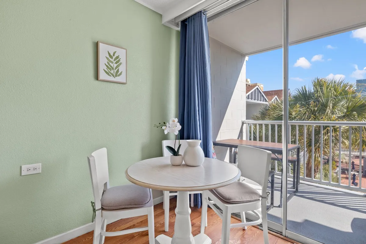 A cozy dining nook inside the condo features a small round table with white chairs, a vibrant potted orchid centerpiece, and blue drapes against a fresh green wall. The space is bathed in soft natural light, ideal for intimate meals, morning coffee, or reading sessions—suggesting comfort and homeliness for longer stays.