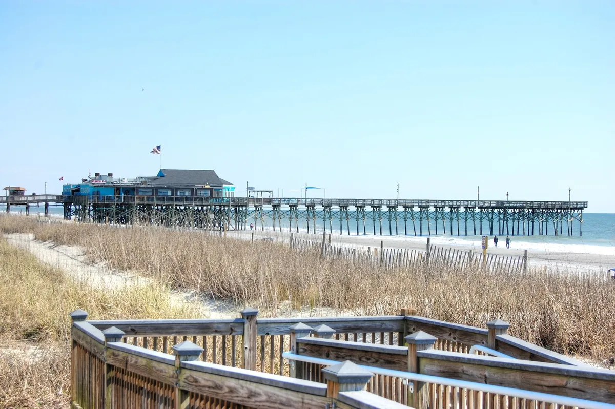 This image captures a tranquil beachfront scene featuring a pier stretching over the ocean, gently rolling sand dunes, and flags fluttering in the breeze. The open sandy coastline invites peaceful strolls, and the distant pier offers a classic touchpoint for sightseeing and fishing. The setting is calm, with soft lighting evoking the off-season serenity of Myrtle Beach.