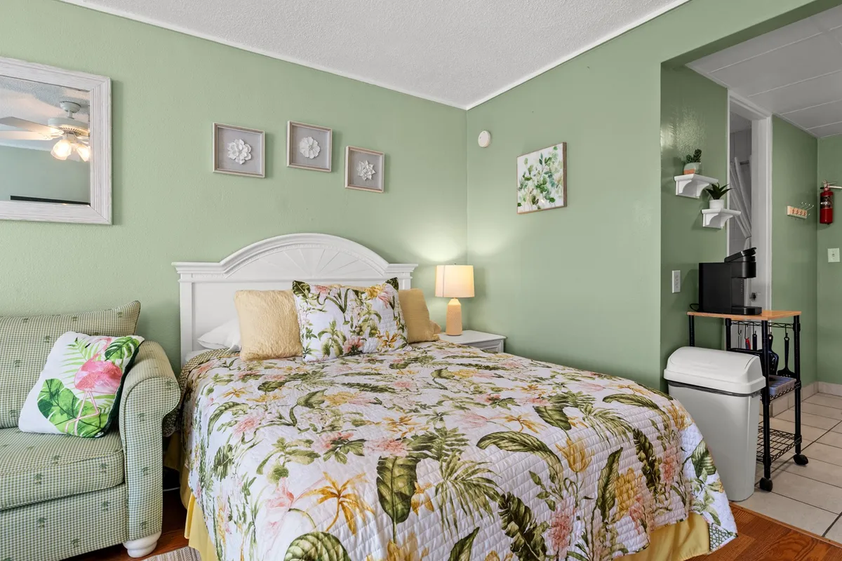 Here, the bed with a floral quilt is bathed in natural light from a nearby window, complemented by a green accent chair and decorative wall art. The bright and cheerful touch, along with the cozy setup, spotlights the restful nature of the sleeping area. The fresh ambiance makes it appealing for both short stays and longer winter escapes.