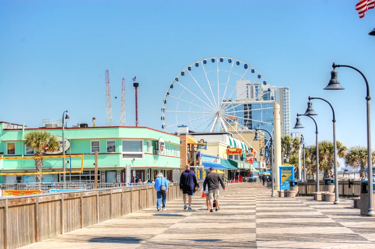 This image captures Myrtle Beach's iconic boardwalk, featuring the vibrant ferris wheel, colorful buildings, and people enjoying a lively stroll. The scene is bustling with activity, highlighting the joyful, family-friendly atmosphere that makes Myrtle Beach a top destination for fun and adventure.