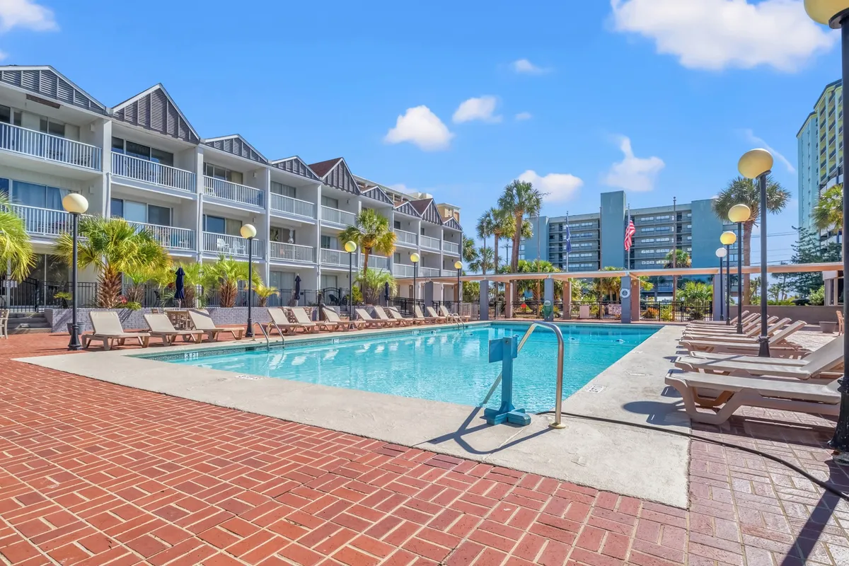 A vibrant outdoor pool scene with inviting blue water, surrounded by comfortable lounge chairs and tall palm trees. The backdrop includes the condo building, hinting at convenient access from residences. The sun is bright, suggesting a perfect day for swimming or relaxing poolside.
