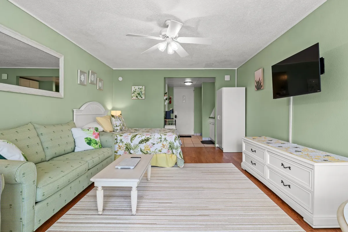 A bright living room scene with a plush sofa, bed, TV, and coffee table, accented by cheerful decorative art. The space feels airy and inviting, perfect for movie nights or daytime relaxation, with ample seating for guests.