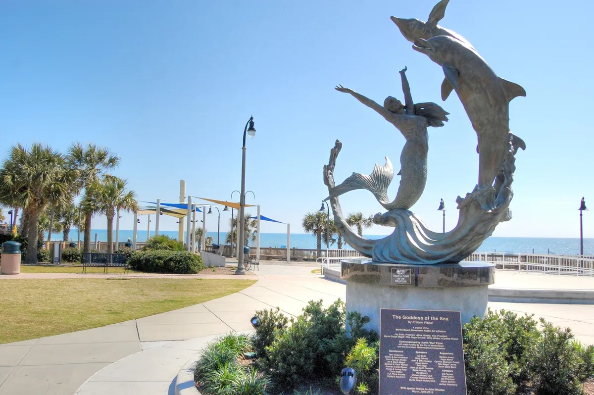 This uplifting scene features the striking Goddess of the Sea sculpture set along a palm-lined promenade with sweeping ocean views. People can be seen strolling, taking in the art and scenery, emphasizing relaxation and memory-making in Myrtle Beach’s beautiful outdoor spaces.