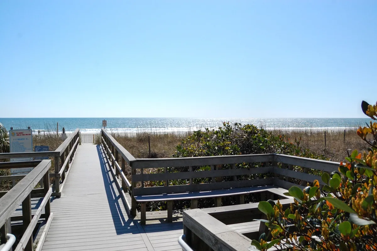 A wooden boardwalk stretches out toward the ocean, bordered by dune vegetation and leading to a panoramic ocean view. The natural setting conveys easy beach access and the allure of the shoreline.