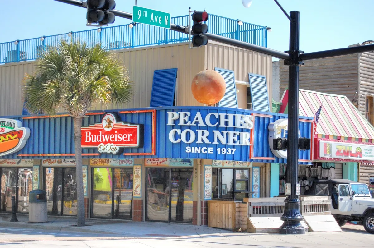 This vibrant exterior shot features the iconic Peaches Corner sign alongside a prominent Budweiser sign, a street sign for 9th Ave N, and festive orange sphere decor. The area is bustling, suggesting an atmosphere filled with dining, socializing, and local flavor.